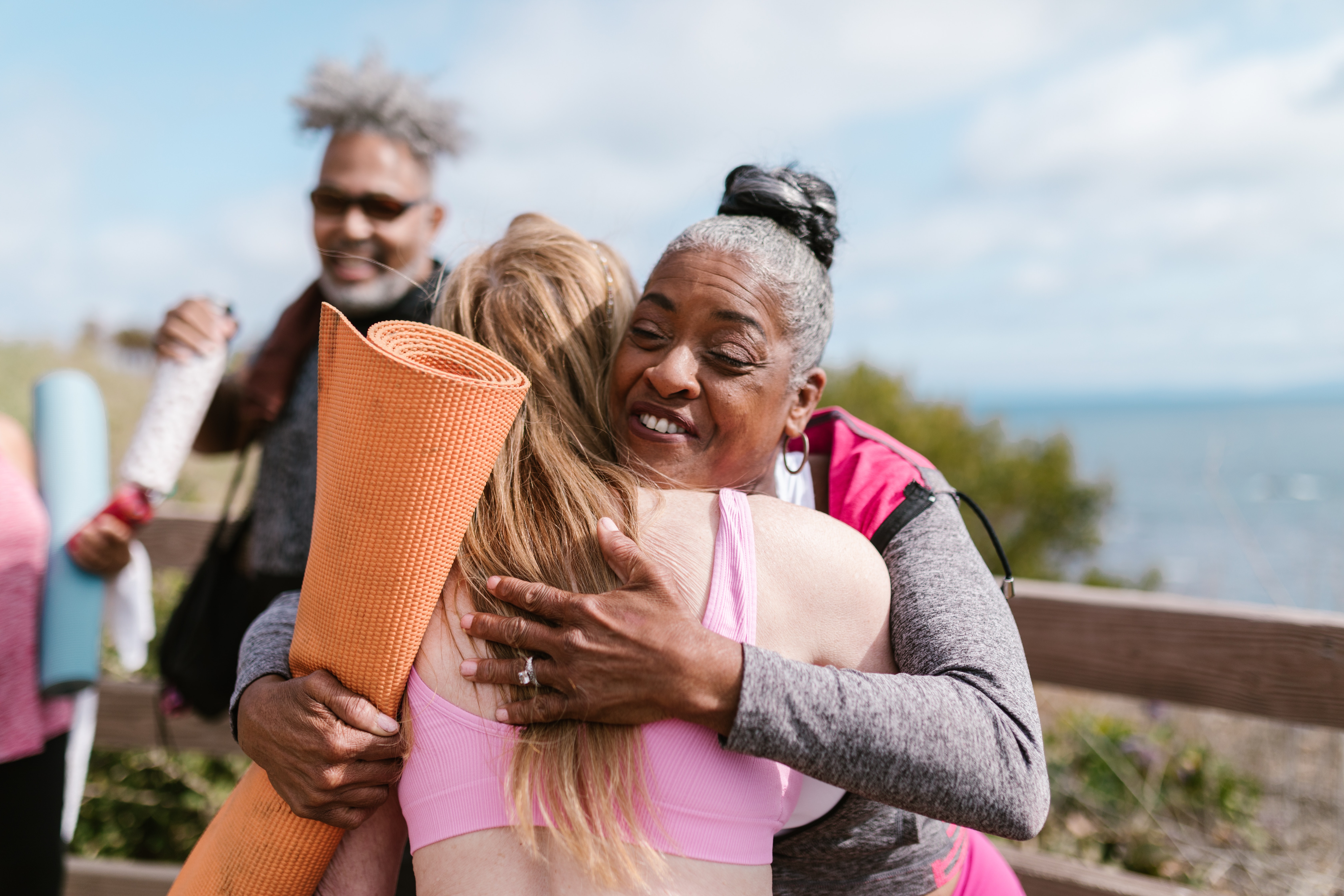 Two people hugging, one with a yoga mat in hand. Ocean in background.